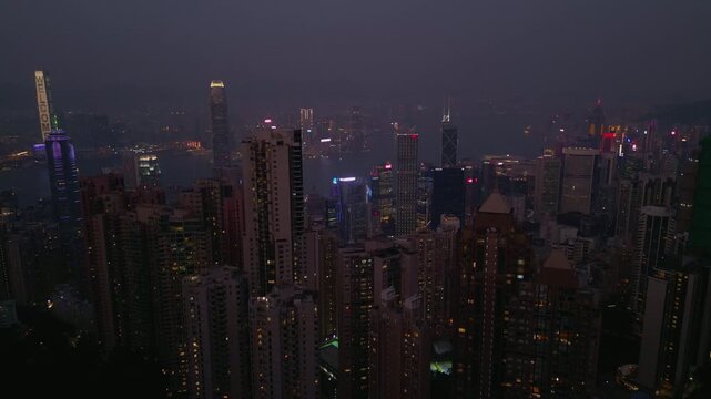 Victoria Peak lookout shows night skyline over harbour haze. Neon signs sparkle on towers and ferry wakes draw thin lines across water as hills fade and windows glow in Hong Kong