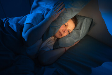 Middle-aged woman lying in bed covering ears with pillows due to noisy neighbors, stressed female...
