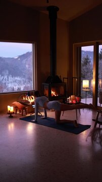 Woman practicing gentle yoga near a fireplace in a cozy mountain house during winter. Warm candlelight, calm atmosphere, slow living lifestyle, evening relaxation indoors