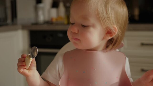 Close-up of a small toddler girl sitting in a high chair at home, licking a spoon and carefully looking at it while eating. Natural everyday moment of child self-feeding, exploring food textures