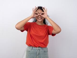 Surprised and excited Asian woman in an orange shirt looking through an OK hand gesture like binoculars, smiling with an impressed and amused expression.