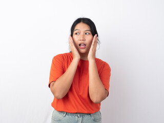 Surprised young Asian woman wearing an orange shirt, hands on cheeks, looking up at copy space, isolated on a pink background.