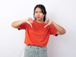 A disappointed Asian woman wearing orange shirt gives thumbs down hand gesture of disapproval, isolated by white background
