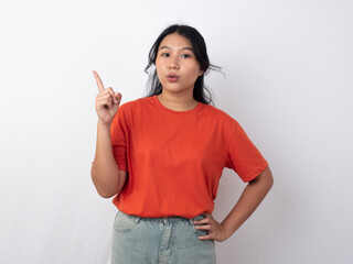 Attractive young Asian woman in orange shirt pointing to the copy space behind her, isolated by white background.