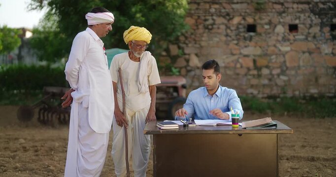 Indian young adult man sitting on chair do visit field work look file check report data at rural area place Banker staff talk speak use ink pad help old aged male to sign thumb print on bank paper