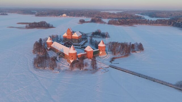 4K aerial drone ascent revealing Trakai Island Castle on Lake Galvė, Lithuania, surrounded by snow and frozen ice at pastel winter sunset, cinematic establishing shot