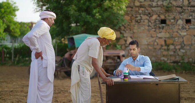 Indian young adult worker staff sitting on chair talk speak share bank loan enquiry old aged farmer at rural area. Banker male do visit field work give cash money to retired man outdoor ground place