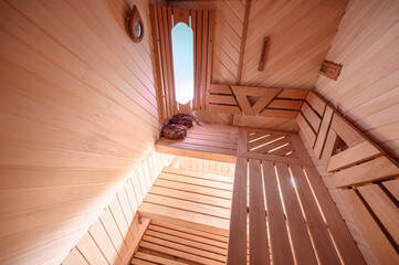 Light wooden sauna interior with slatted benches, oval window, heated stones on upper shelf, wall clock, warm natural finish, cozy and functional design.