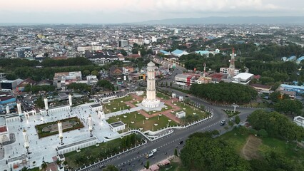Aerial view of a grand mosque and monument in a city surrounded by lush greenery and modern buildings with tall minarets and a prominent white tower.