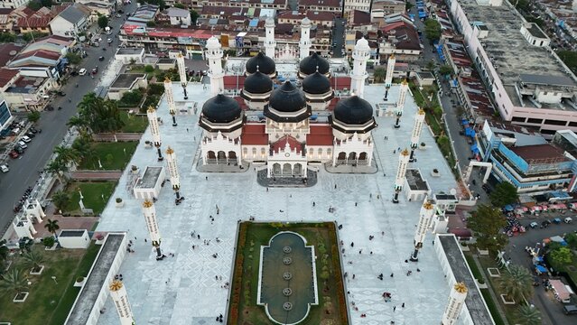 Aerial view of grand mosque, Masjid Raya Baiturrahman, Banda Aceh, Indonesia, with multiple black domes and white minarets in an urban setting surrounded by buildings and streets, with people walking 