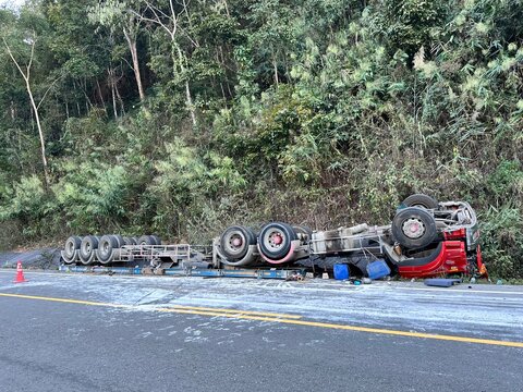 A high-impact shot showing a large red semi-trailer truck completely overturned on the side of a winding mountain road.