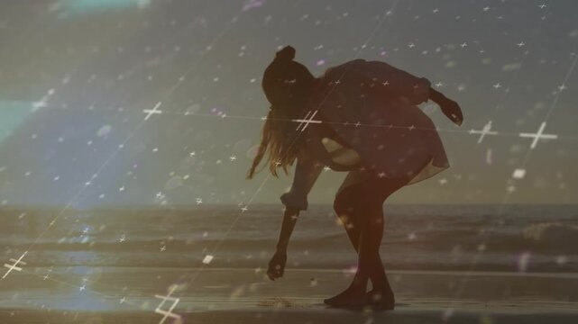 Woman walking wet sand, spotting shell and bending to pick up and study it, film-grain overlaying