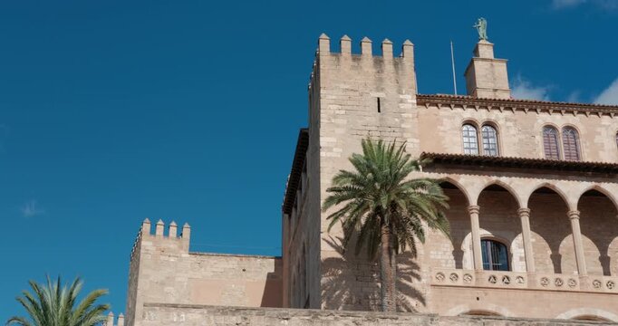 Royal Palace of Almudaina stands under a bright blue sky in Palma de Mallorca, Spain. Historic architecture with palm trees on a clear day.