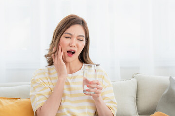 Fototapeta premium Young Asian woman at home holding a glass of water and touching her cheek with an expression of sharp pain or discomfort. Concept of sensitive teeth, toothache, and dental problems.