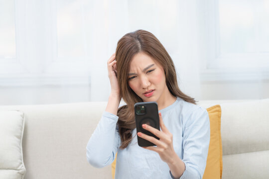 Stressed young Asian woman scratching her head while looking at a suspicious or confusing message on her smartphone at home. Concept of online scams, debt, and digital fraud.