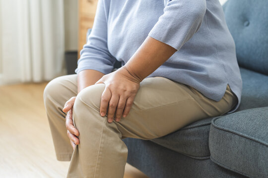 Body arthritis muscles stiff problem, aged woman suffering on leg, pain knee from disease chronic, hand massage rubbing joint injury, mature hurt or sore sitting on sofa at home. Health care people.