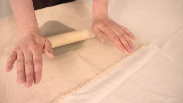 Close up footage of a ceramic workshop where an artisan rolls a soft clay slab using a wooden rolling pin. Traditional pottery process showing hands shaping natural clay with simple tools 