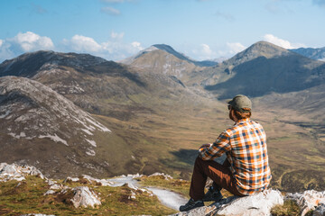 Fototapeta premium A hiker sits atop a rugged quartzite peak in Connemara National Park, surveying the dramatic Twelve Bens mountain range under a vast, cloud-filled sky.