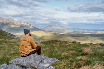 Fototapeta premium A hiker sits atop a rugged peak in Connemara National Park, surveying the beautiful Irish landscape under a vast, cloud-filled sky.
