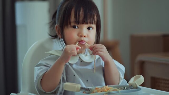 Cute Asian toddler girl eating spaghetti with messy face on high chair. Childhood nutrition self-feeding and healthy development concept. Little girl enjoying mealtime at home