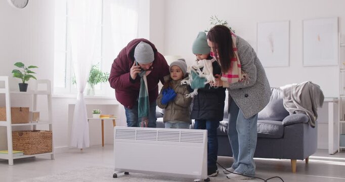 Frozen family with kids in winter outwear standing near electric heater at home warning up and calling public utility service by mobile phone. Heating problems. 4k video. Slow motion video.