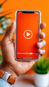 A hand holds a smartphone displaying a play button on a red screen. The background is orange with a small plant beside it. This scene shows a casual moment