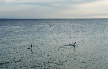 A calm blue sea with two surfers walking into the distance
