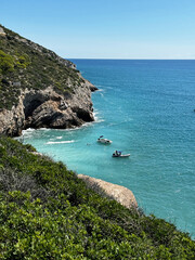 An azure wild beach with black rocks, an inaccessible shore with approaching boats.