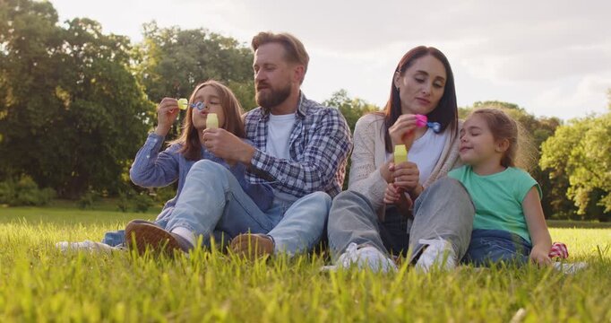 Summer fun holiday family sitting on green park lawn, blowing soap bubbles playing, enjoying outdoor leisure time, parents and kids engaging in activities for amusement, relaxing enjoyment weekend