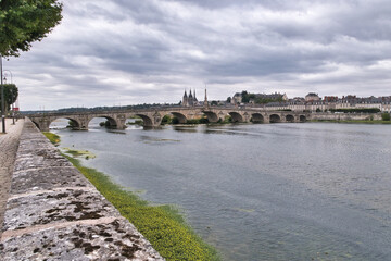 Obraz premium Pont Wilson Bridge and Blois Skyline - Wide view of the Loire River with the historic Pont Wilson stone bridge and the city skyline of Blois, France, under a cloudy sky.