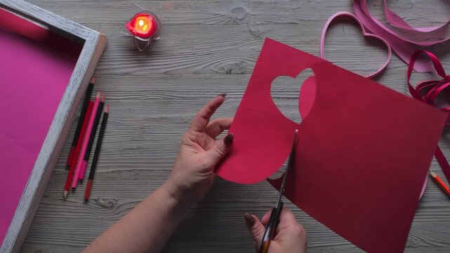 Top view of hand cutting heart shapes from red cardboard with scissors. DIY handmade Valentine Day card preparation and creative craft process