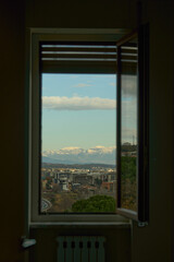 Fototapeta premium Open window showing city landscape, snow capped mountains