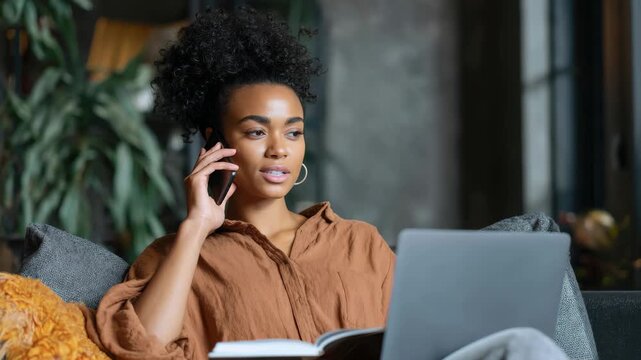 Black woman sits on couch reading a book while on phone call using laptop in cozy living room during daytime hours
