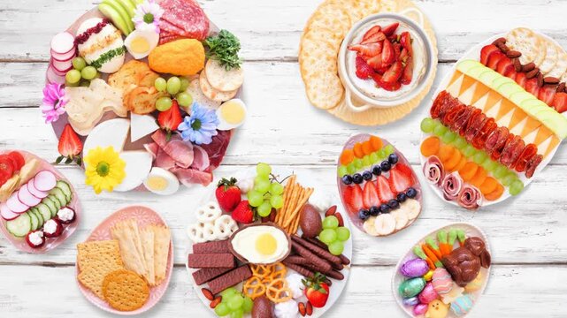 Spring or Easter grazing board theme table scene against a white wood background. Stop motion table placement. Overhead view. Assortment of sweets and cheese, meat and fruit appetizers.