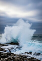 Naklejka premium Turbulent, cold blue ocean water crashes forcefully against rugged, dark coastal rocks, creating dramatic white spray and sea foam under a moody, cloudy sky, liquid, environment, texture