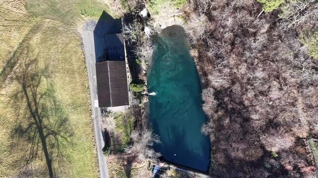 Top-down aerial drone footage rising above a small river dam, revealing water inflow and outflow, nearby building, and surrounding trees in a rural landscape.
