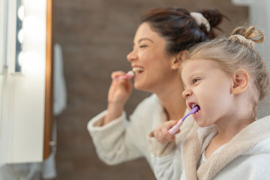 Smiling mother and young daughter in bathrobes brush their teeth together in a bathroom mirror, showing daily hygiene routine, positive parenting, learning by example, and warm family bonding at home.