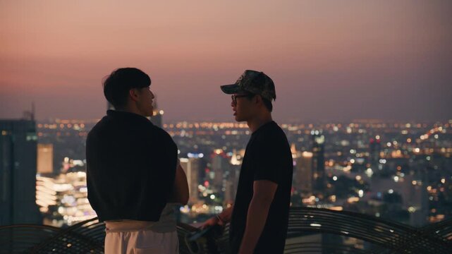 Two asian men chat on top of a high rise building, enjoying the panoramic terrace view of lights of Bangkok as the sun sets