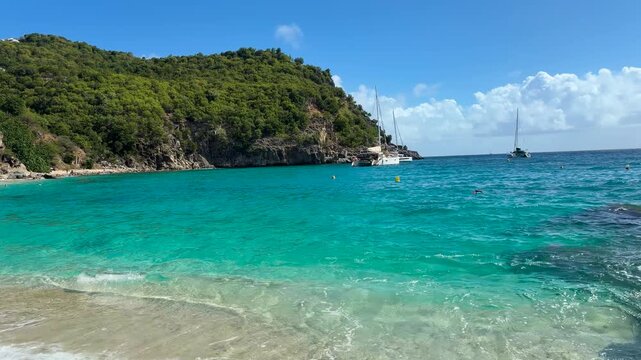 A sandy shore with turquoise blue water overlooks lush cliffs and moored sailboats on the Island Saint Martin, the Caribbean.