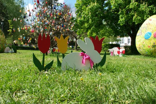 Colorful Easter decorations in a city park during springtime. A large hand-painted Easter egg and an egg tree stands among decorative easter bunny and tulip ornaments on green grass. Open copy Space.