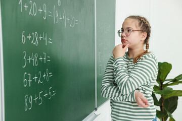 Thoughtful schoolgirl near chalkboard with math assignment in classroom