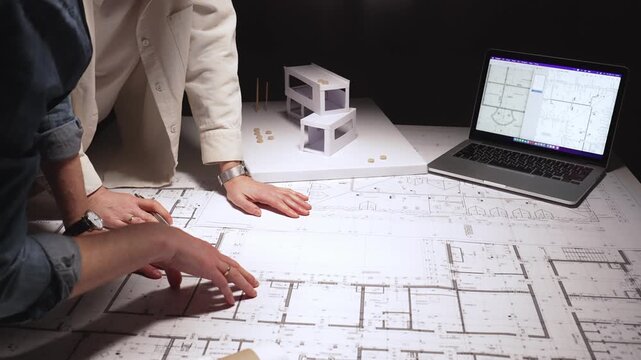 Architects collaborating on building plans with a laptop, reviewing blueprints and architectural models in a modern design studio, discussing future construction projects