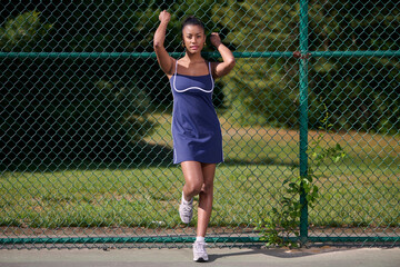 Obraz premium Beautiful African-American female tennis player in blue dress standing at fence - holding fence behind her