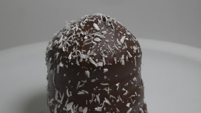 A close-up shot of a glazed dessert topped with crushed nut pieces, seen slowly rotating against a clean, neutral background.