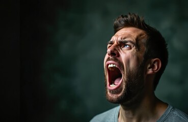 Fototapeta premium Man with beard screams intensely, mouth wide open in rage. Extreme emotion on face, head tilted back, eyes looking up. Dark background emphasizes his loud outburst and frustration.
