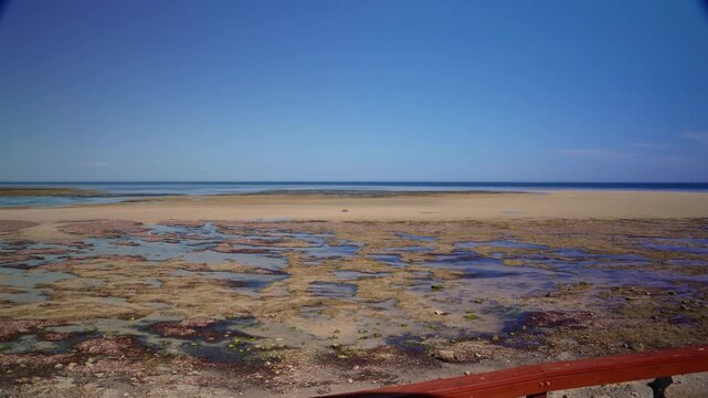 A 45 second timelapse reveals a beach at low tide featuring a coral reef and sandbar under a clear blue sky, as the water rushes in to fill all of the exposed area and it begins to get dark.