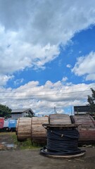 Large wooden cable reels and coiled black wires sit in an open industrial yard under a bright blue sky, blending urban infrastructure with dramatic clouds.