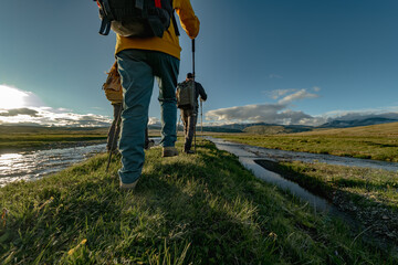 Obraz premium Two hikers with backpacks and trekking poles are walking along a narrow grassy ridge beside a river in a vast, green mountain landscape under a bright, sunny sky.
