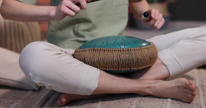 Woman playing steel tongue drum with mallets on floor at home, closeup