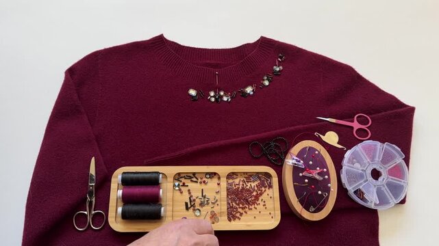 Time lapse video of woman's hand using tweezers to arrange beads and bugle beads on burgundy sweater before sewing them on. Handmade decorating women clothing. Close-up of sew accessories